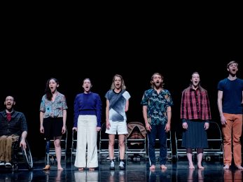 7 dancers stand in a line at the front of the stage with mouths open as if shouting