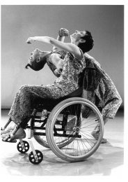 one dancer in a wheelchair leans back with another dancer standing behind them leaning back too.