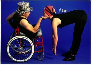 one male dancer is in a wheelchair wearing plain black clothing and a top hat with his right hand out. Resting on that hand is a female dancer also wearing plain black clothing but wit a red knitted hat. Both are looking at each other in the eye.