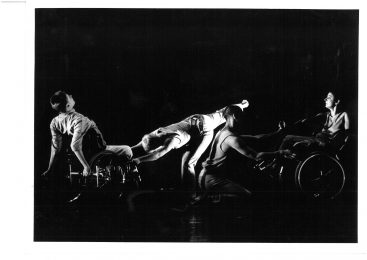 4 dancers are on a dark-lit stage. 2 duets are on the stage. One involves a a man leaning back in a wheelchair with the other dancer tilting down and holding on to the front of the wheelchair. The other duet involves one sitting in a wheelchair and the other crouching down, both looking at each other, holding hands.