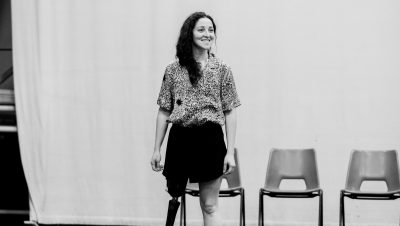 a dancer stands in front of a row of chairs looking to her left, smiling.
