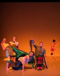 Disabled and non-disabled dancers pull on each other's clothes, one lifts a long-haired wig