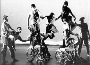 A black-and-white photograph of a group of dancers, including wheelchair users, forming interconnected balances and lifts across the stage, creating a dynamic sense of teamwork and movement.