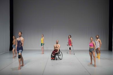 Disabled and non disabled dancers stand on a white stage with bright coloured clothing. All stand facing the diagonal, looking straight ahead.