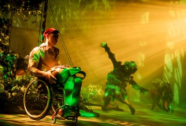 Jungle scene with creature in background reaching up from knees. Boy using wheelchair downstage looks amazed.