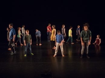 A large group of people stand spread out on a dark stage. They all face different directions with some of them having their eyes closed.