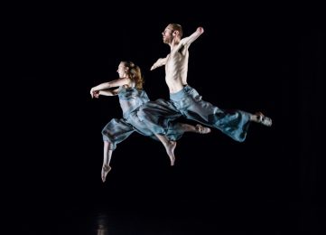 2 dancers are in mid with their right legs bent and their left legs straight behind them. They both look ahead with one dancers arms open out and the other with their arms curved in front of them.