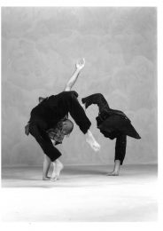 2 male dancers in plain clothing have their weight on one of their hands with their bodies raised up in the air. Their backs are away from the camera.