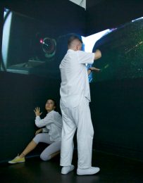 Two dancers, one standing and one kneeling interact with projection on dark wall
