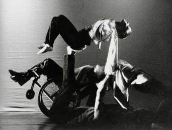 A black-and-white photograph of dancers creating an intertwined lifting shape, with a wheelchair incorporated into the movement, conveying strength, balance and collaboration.