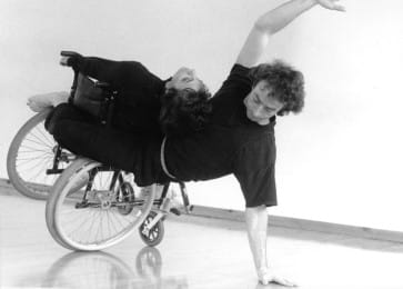 A black-and-white photograph of two dancers balancing together, one using a wheelchair, creating a fluid and connected movement pose.