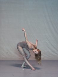 Dancer wearing a skin coloured leotard bends forward with her right leg stretched out and arms extended behind her.