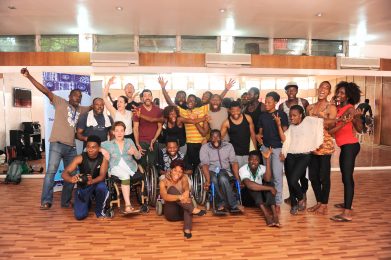 A group of disabled and non disabled dancers stand close together looking at the camera smiling, some have their arms in the air.