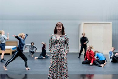 a woman stands in front of a stage. Behind her are dancers in mid-movement