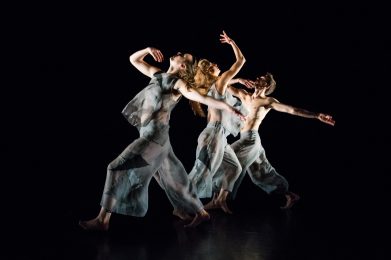 three dancers stand in a diagonal line facing different directions. All of them are looking up with their right hand below their chin and the left hand stretched out.