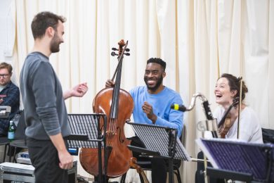 2 people are seated, one with a cello and another with a violin. They are both smiling at someone who is standing up, looking at them.