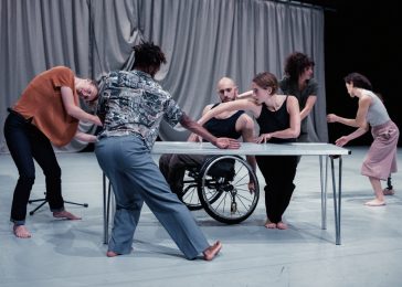 People move around a grey table arms overlapping with arms, reaching around each other. There is a grey draped curtain in the background and two other people facing each other mid movement.