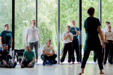 A led Artist teaching a class to a group of young participants in a bright studio space. The photo is captured from the teacher's back and the students are standing in front of a large glass window.