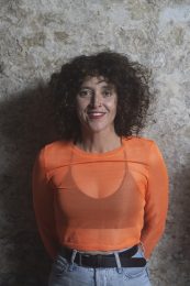 Headshot of Anna Seymour, smiling with short curly brown hair. They are wearing an orange top with a black bra visible underneath. Anna self-describes as: a white woman in her 40s with dark curly hair that comes to just above the shoulder and a wavy fringe. They have a nose ring and freckles. They are Deaf and have a petite frame.