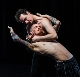 2 men embracing. One man has his hands gently wrapped around the neck of the other. The other man is looking away from him but leaning back on him. There is a comfortable energy between the pair.