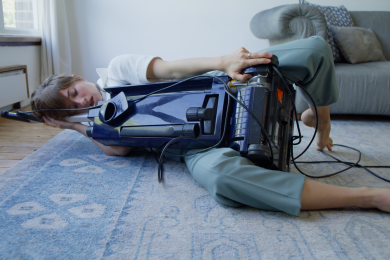 A white woman lies on her side in a living room, embracing a dark blue upright vacuum cleaner, one leg wrapped around its wheeled base, her bare foot arching against a pale blue patterned carpet. She cradles her head against the handle of the cleaner, echoing the soft curve of a greyish blue sofa behind.