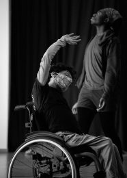 Kimberley reaches her front arm up and over her head with palm facing up, her head is tilted in the same direction as her arm. She is using a wheelchair. Another dancer walks behind her.