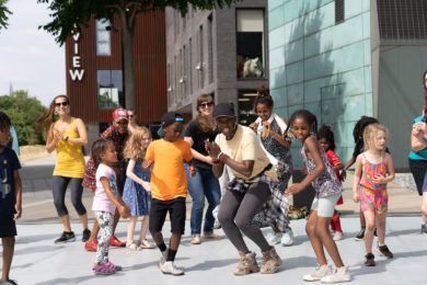 A group of young people taking part in the Afro Fusion workshop at Candoco's Day of Dance 2022. They are dancing outdoors on a large dance mat located in front of the Mountview building, in Peckham Square. The participants wear bright colourful clothes and are following the steps of the choreographer Fumi Opeyemi.