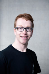 Dermot Farrell. A smiling headshot of a dancer with short red hair and black framed glasses. He is wearing a black tshirt.