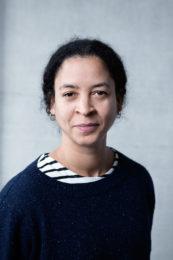 Vanessa Abreu. A smiling woman with dark hair is wearing a dark blue jumper. We can just see a white tshirt with black strips appearing at the neckline