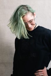 A person with light green hair and a black top is smiling and looking down.