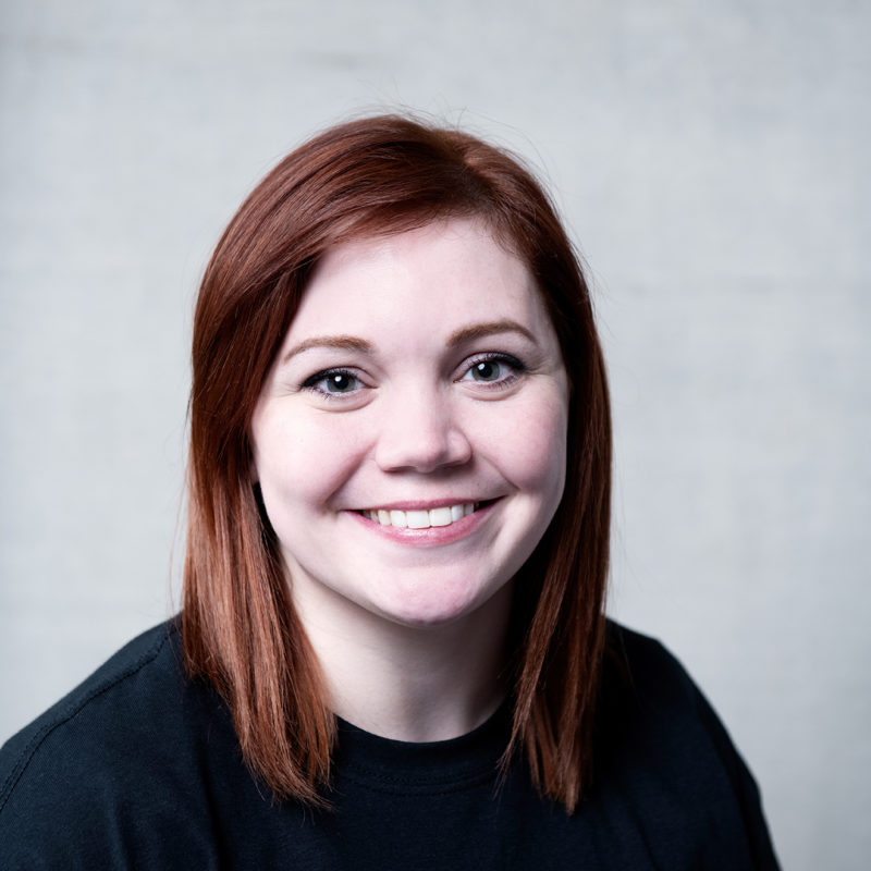 Hannah Raynor. A headshot of a smiling person with long reddish brown hair who is wearing a black t-shirt
