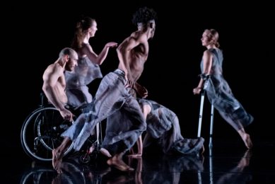 Five dancers moving quickly around the stage. They wear sheer, light grey, loose clothing and look toward each other as they move.