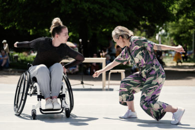 Two dancers, Maiya Leeke and Caroline Lofthouse performing 'I think we should start over' outdoors in front of a live audience. Maiya identifies as white woman in her mid 20s, she has blonde hair, tied up into a messy bun and is wearing a black top and grey trousers. Caroline also identifies as a blonde, white woman and she is wearing a pink and green jumpsuit. Maiya is bent forward with one arm stretched backwards and one hand on her wheelchair. Caroline is also bent forward on one knee and has both of her arms open with her fists clenched.