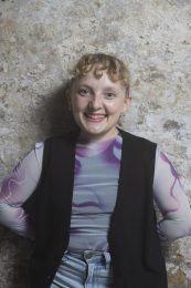 Headshot of Annie Edwards, a person with short, blonde curly hair, smiling at the camera. They are wearing a grey and purple top layered under a black gilet jacket, with their arms positioned behind their back. Annie self-describes as: a small white woman in her 20's with short curly hair.