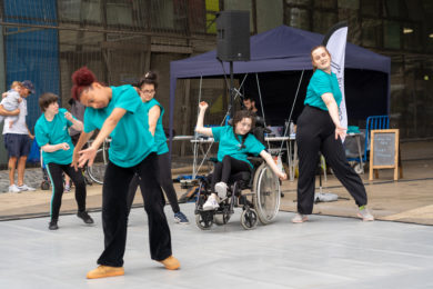 Participants dancing outdoors at annual Day of Dance Festival 2022. They wear teal t-shirts and black trousers. They are standing on a dance mat and appear to be are moving towards different directions.