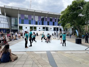 Group of dancers in teal t-shirts getting ready for the start of their performance. They are in Peckham Library Square, on a grey dance floor. There are around 40 audience members watching.