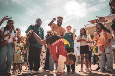A group of people standing in a circle around a child who is doing a headstand. The crowd is cheering and smiling whilst watching the dancing.
