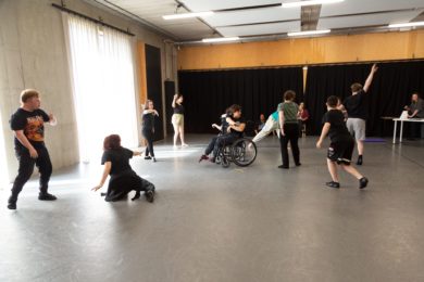 A group of around 10 dancers, rehearsing in a light studio space.