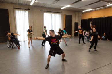a group of around 15 dancers all doing different movement shapes. There is one dancer in the foreground in a low squat position with his arms out horizontally.