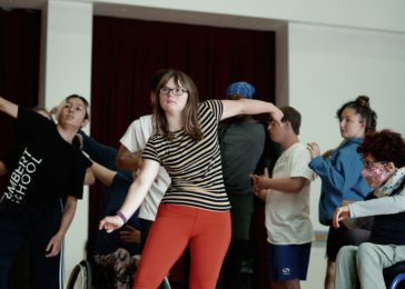 A group of dancers looking towards the left. One dancer in the foreground has bright coral coloured leggings and a black and brown striped top. They wear with glasses and have light brown shoulder length hair.