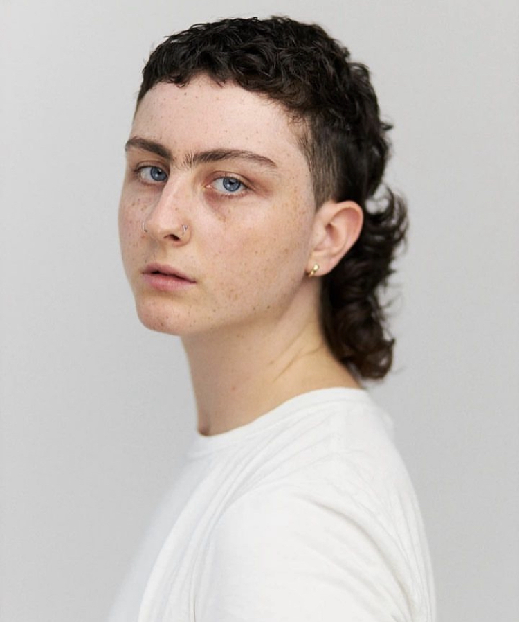 Bel is looking direction at the camera with their body facing the left side of the frame. They are posing in front of a grey background. Bel has short brown hair and it wearing a plain white tshirt.