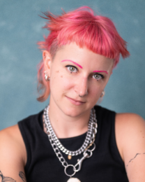 Jay is looking into the camera with a blue background behind them. They have short pink hair and they are wearing a black vest top with some silver necklaces.