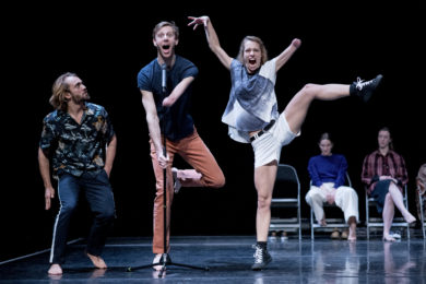 Five dancers on stage, two are sat at the back and three are lined up at the front. The person in the middle is standing in front of microphone with their mouth open and a surprised look on their face. On the right, one person making a funny face with arms raised above their head and leg raised off the ground. The person on the left is looking at the dancer in the middle with a serious look on their face.
