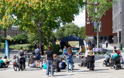 Audience, seen from the back, watching a outdoor duet. In the background there is a brown building with a sign reading 'Mountview'.