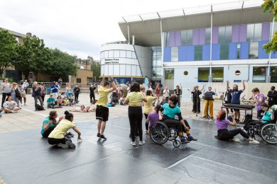 A group of people wearing yellow, purple and teal coloured t-shirts are dancing outdoors on a large grey dance mat. A live audience is cheering and clapping to the performance.