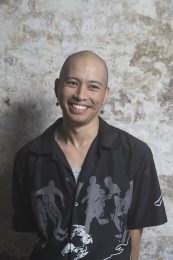 Headshot of James Olivo, a person with a shaved bald head and wearing earrings and a black long-sleeve shirt. They are smiling and looking slightly to the side, away from the camera. James self-describes as: a Filipino-Chinese person in their late 20s, with a shaved head.