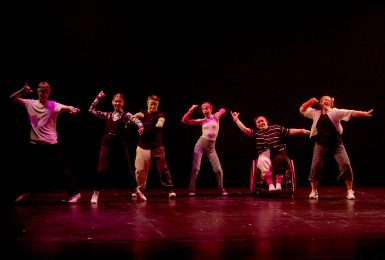 Six young dancers wearing brightly coloured armbands and headbands performing on stage raising their right arm. One dancer is using a wheelchair.