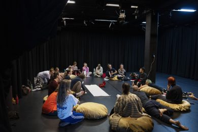 A large group of people, sitting in a circle in a dimly lit indoor space. Some are sitting on bean bags whilst others are lying down. White paper sheets are spread on the floor, along with a box of pens for use during the workshop.