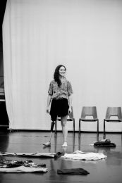A black-and-white photo of a person wearing a prosthetic leg, with long, dark, hair styled to one side. She smiles warmly while looking away from the camera. She stands in a space with three empty chairs in the background. The floor is scattered with various costumes, and a white curtain hangs in the backdrop, providing a soft, neutral tone to the scene.
