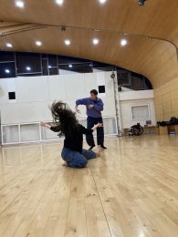 Two dancers in a studio: one with long, flowing hair kneels on the floor, extending one leg forward and arms open. In the background, another dancer stands with one arm extended to the side, elbow slightly bent, and hand pointing downward.